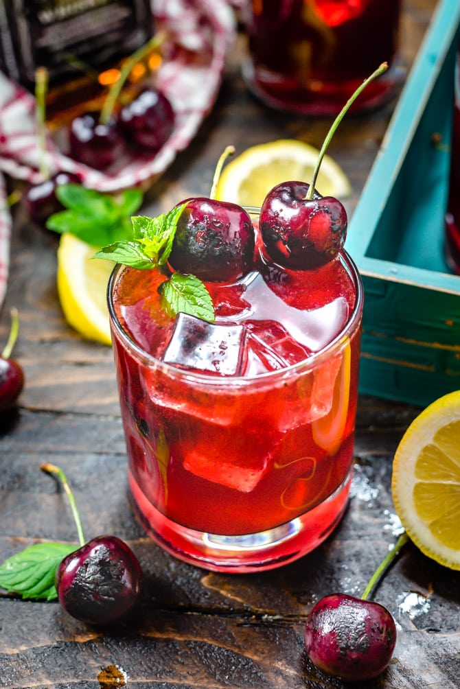 A red Charred Cherry Whiskey Lemonade cocktail in a small glass with mint, ice, and charred cherry garnishes.