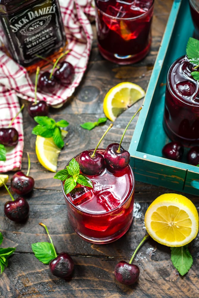 An overhead shot of a tabletop with Charred Cherry Whiskey Lemonade cocktails in small glasses, a Jack Daniels whiskey bottle, fresh mint, charred cherries, and lemon wedges.