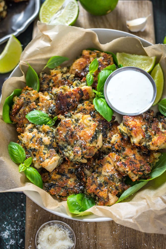 Spinach and artichoke dip chicken fritters in a parchment lined bowl with basil leaves surrounding them, and sour cream in a condiment container.