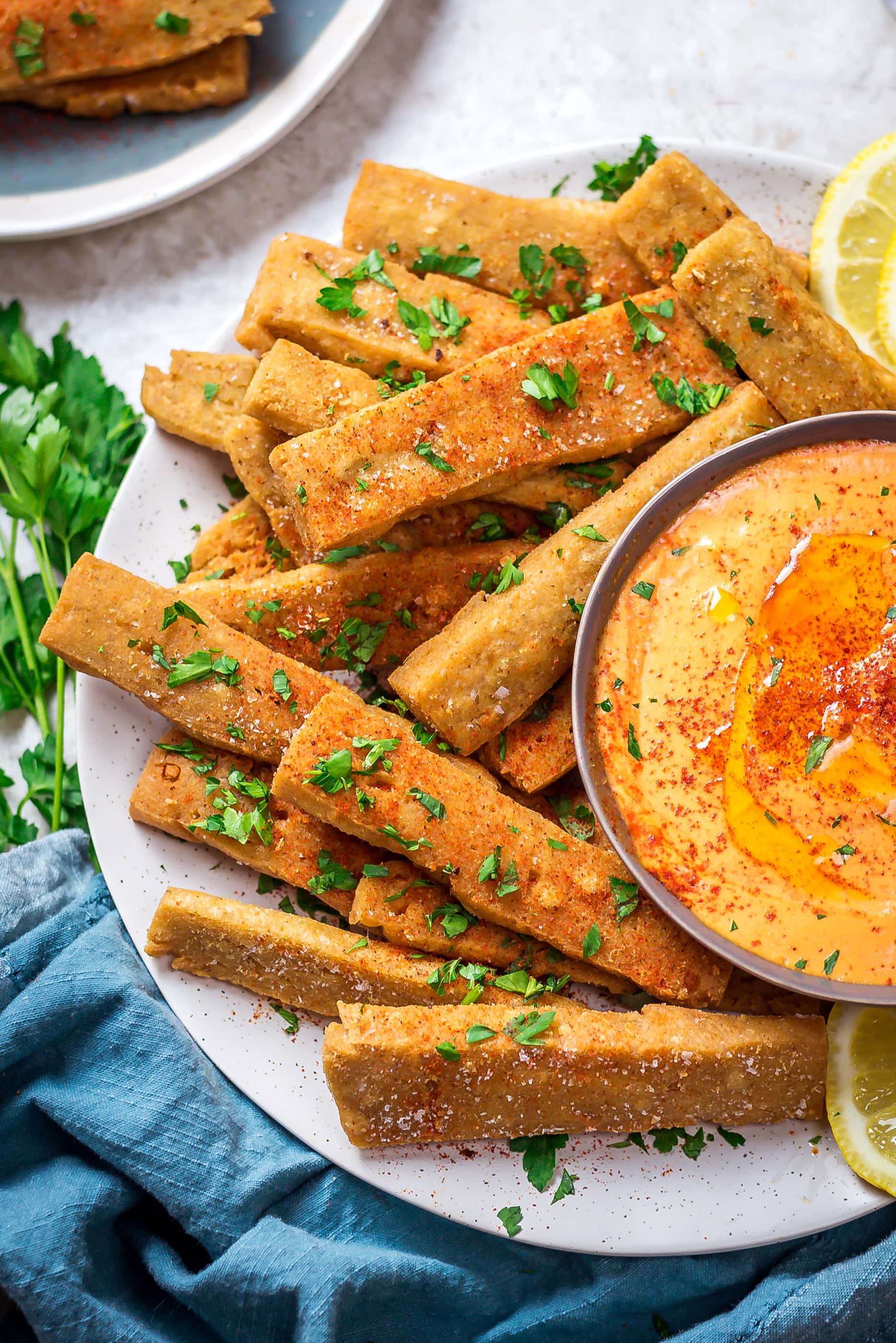 A close up of Hummus Fries with Spicy Tahini Dip on a round white plate with a blue cloth below.