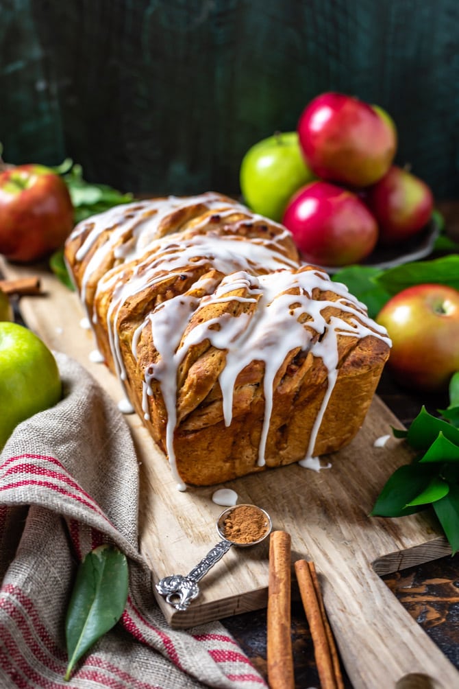 A finished shot of Apple Fritter Pull-Apart Bread on a wooden cutting board, surrounded by apples and cinnamon sticks.