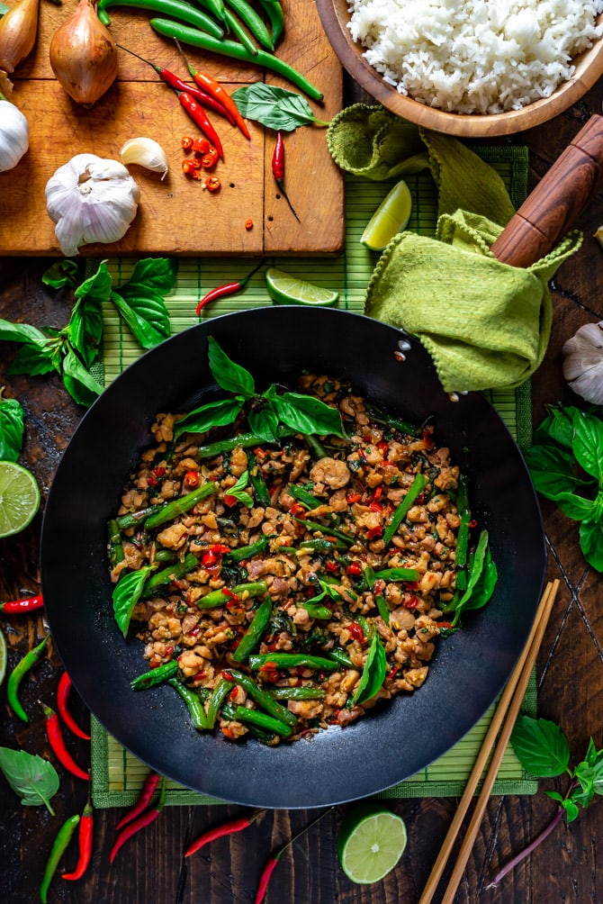 A zoomed out overhead view of 15 Minute Thai Basil Chicken in a wok surrounded by thai basil, limes, and thai chilies.