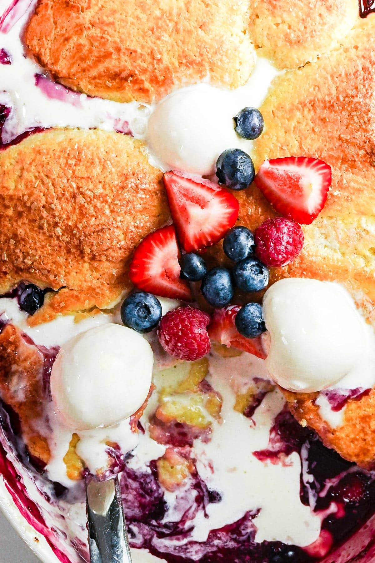 Close-up photo of Berry-Buttermilk Cobbler with blueberries, raspberries, and strawberries on top, and vanilla ice cream.
