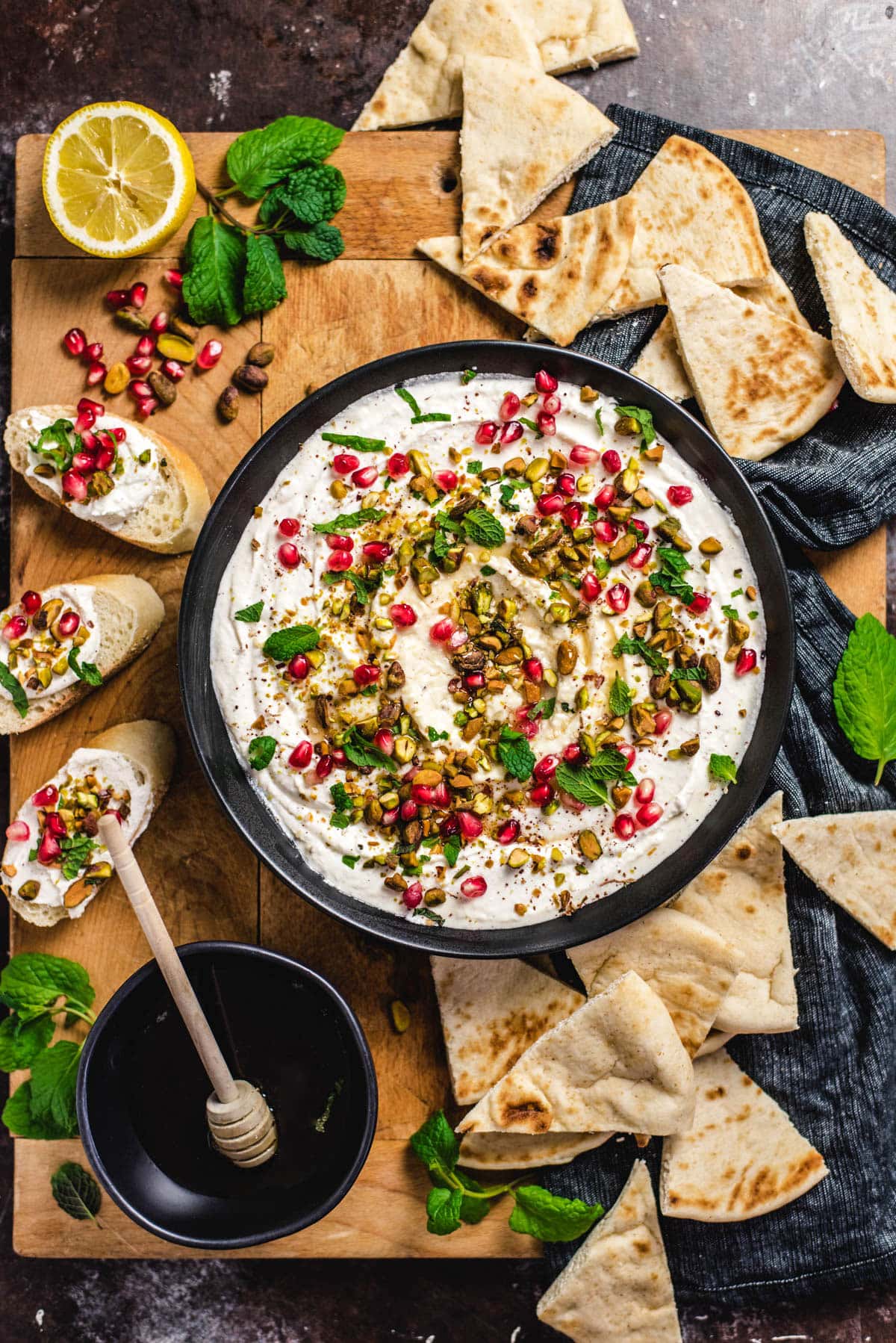 An overhead picture of a bowl of Honey-Whipped Feta Dip. The bowl is black and the dip has been swirled in the bowl and topped with honey, bright red pomegranate seeds, green pistachios, and fresh mint leaves. It's surrounded by pita bread and crusty bread.