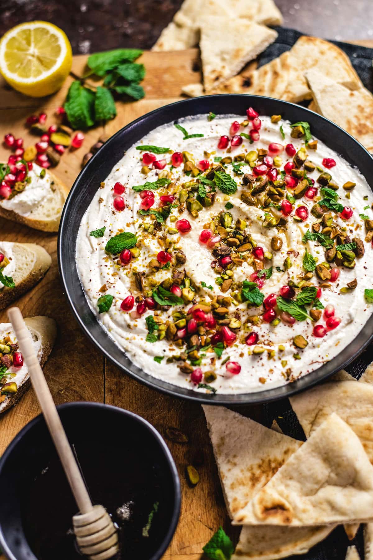 A close-up picture of Honey-Whipped Feta Dip in a black bowl, surrounded by pita triangles and slices of crusty bread. There is a black small bowl of honey in the corner as well as a lemon, mint, and pomegranate seeds scattered about.