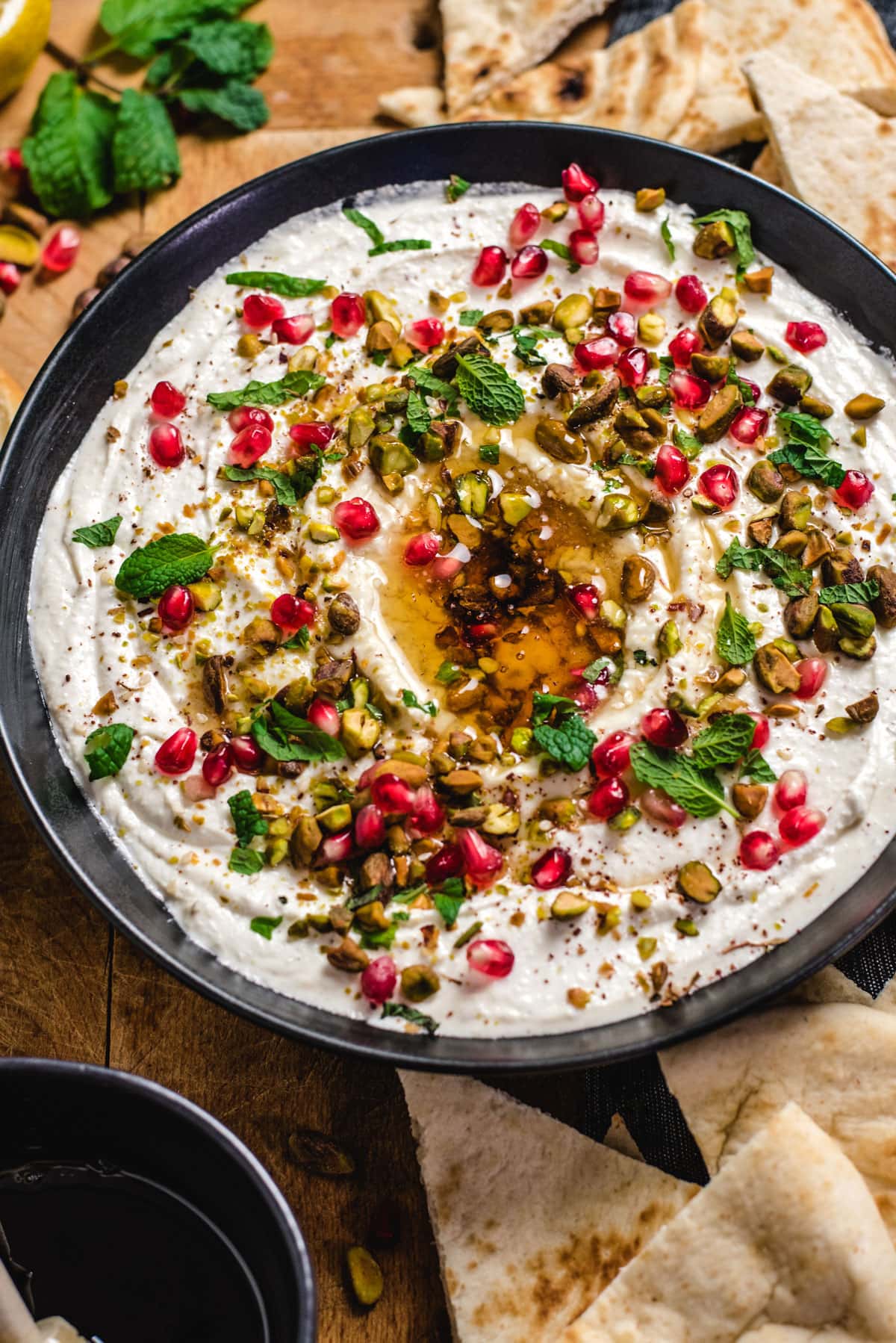 A close-up picture of a bowl of Honey-Whipped Feta Dip. The bowl is black and the dip has been swirled in the bowl and topped with honey, bright red pomegranate seeds, green pistachios, and fresh mint leaves.