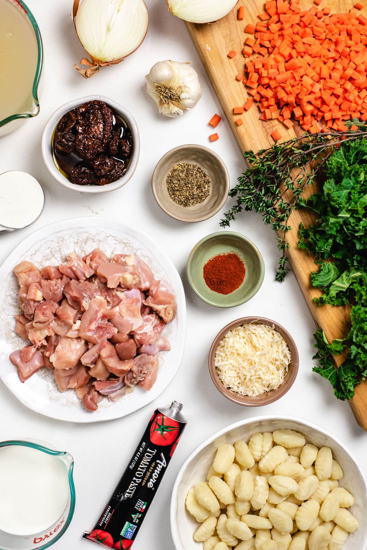 An overhead shot of ingredients used to make chicken gnocchi soup. Pictured in order from top left to right bottom are chicken broth, a halved onion, a head of garlic, chopped carrots, flour, sun-dried tomatoes, Italian seasonings, fresh thyme, chicken thighs, smoked paprika, kale, parmesan cheese, half-and-half, tomato paste, and fresh gnocchi.