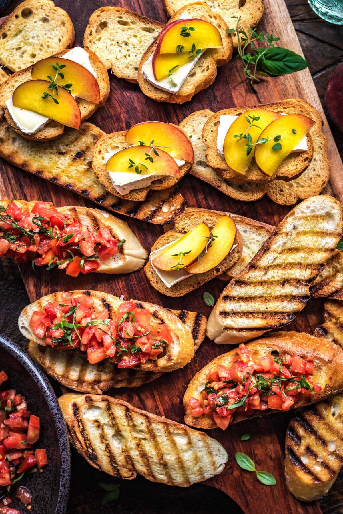 Various grilled and toasted slices of crostini and bruschetta are placed on a diagonally-laid wooden cutting board. Some of the crostini are topped with sliced brie, fresh peach slices, and thyme. Some of the bruschetta toasts are topped with a tomato-basil mixture.