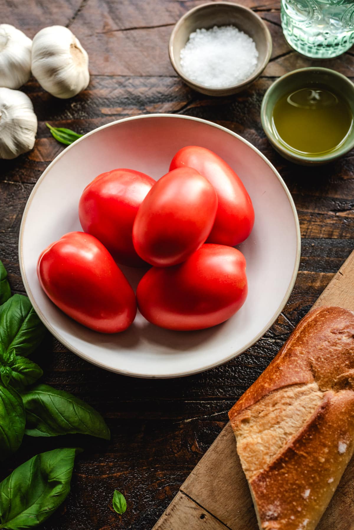 A photo of roma tomatoes, bread, fresh basil, garlic, coarse salt, and olive oil. 