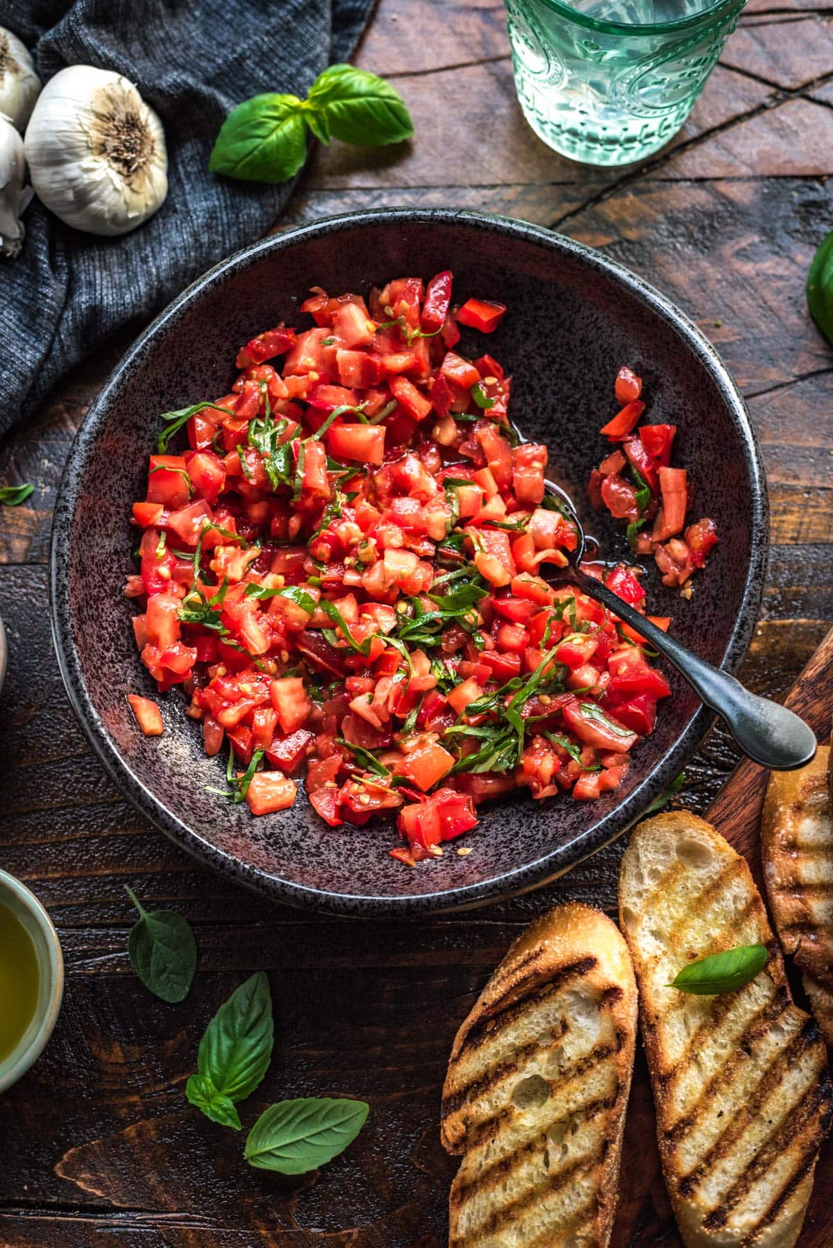 A large black bowl filled with a vibrant chopped tomato and basil mixture, with a spoon set in the mixture, and grilled bread on the side.