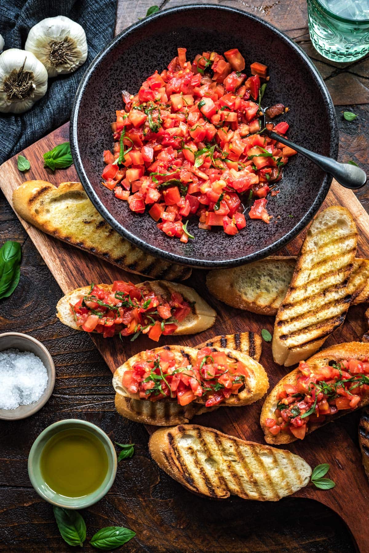 A picture of grilled bread on a cutting board, topped with tomato and basil bruschetta from a large black bowl. 