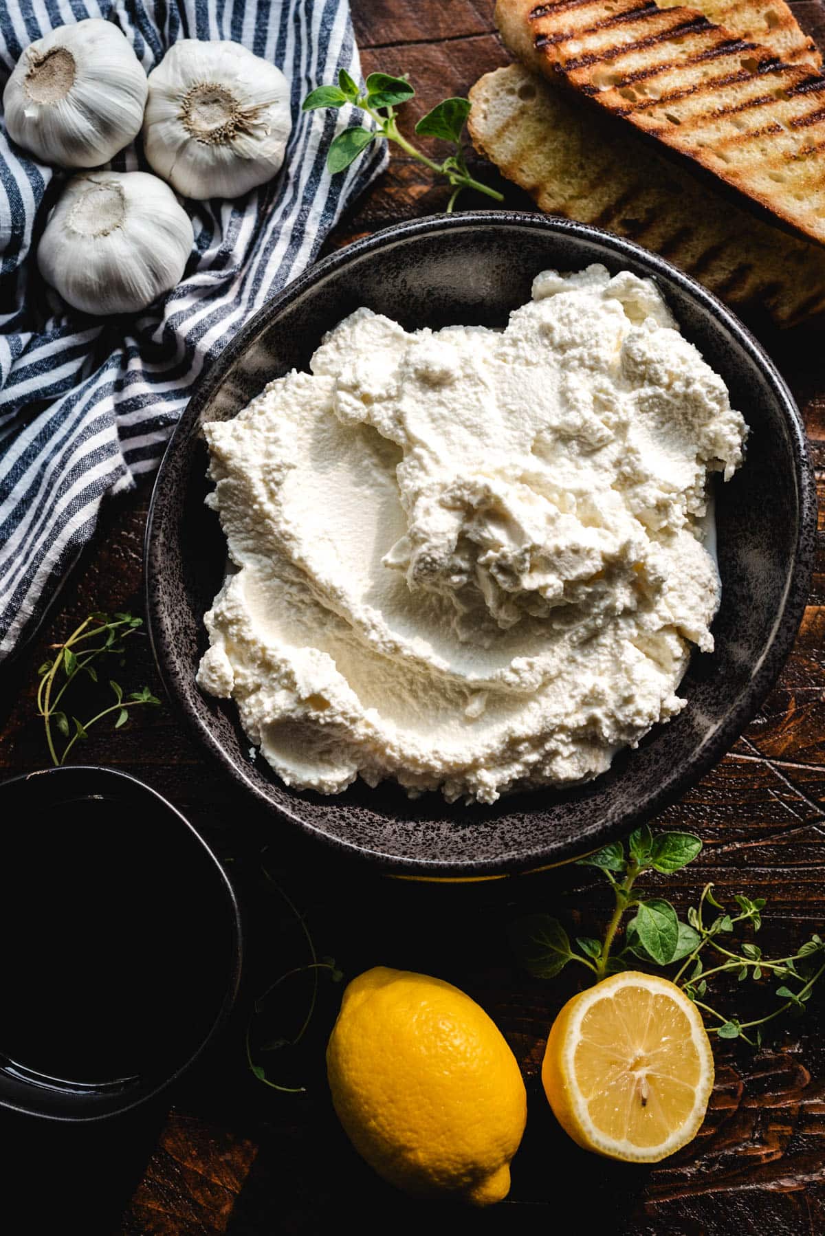 A photo of ingredients for whipped ricotta: ricotta cheese, grilled bread, olive oil, lemons, herbs, and garlic.