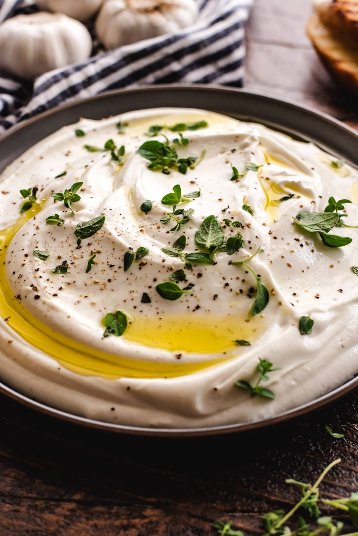 A side view, close up shot of a gray bowl that is full of whipped ricotta. The ricotta is topped with small thyme and oregano leaves, black pepper, and a drizzle of olive oil. 