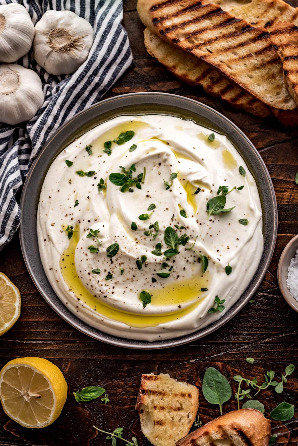 A large gray bowl holds whipped ricotta which is topped with small leaves of fresh oregano and thyme, cracked black pepper, and a drizzle of olive oil. The bowl is on a rustic wooden table, and surrounded by grilled bread, a gray and white striped towel, halved lemons, and a pinch bowl of coarse salt.
