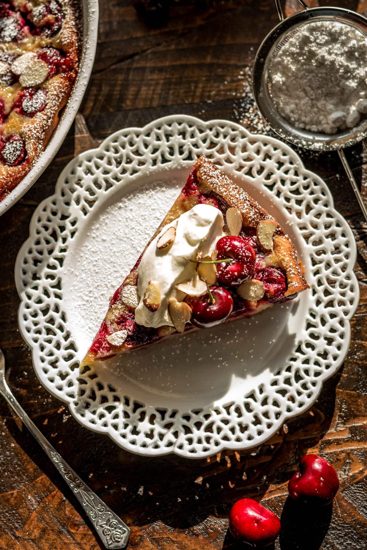 A slice of cherry clafoutis topped with fresh cherries, whipped cream, powdered sugar, and sliced almonds atop a lace-rimmed white plate.