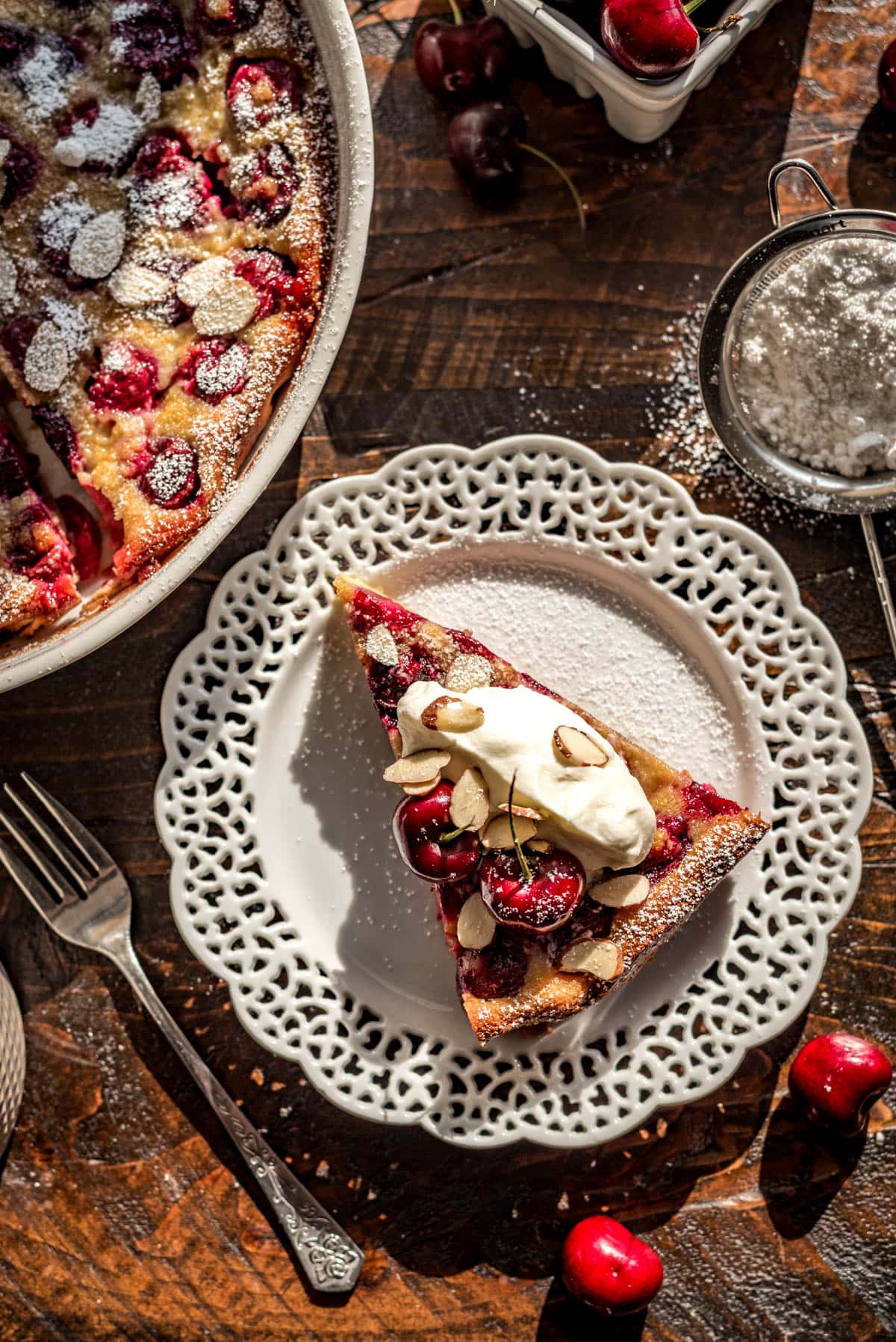 A slice of cherry clafoutis topped with fresh cherries, whipped cream, powdered sugar, and sliced almonds atop a lace-rimmed white plate, with a dish of baked clafoutis in the corner.