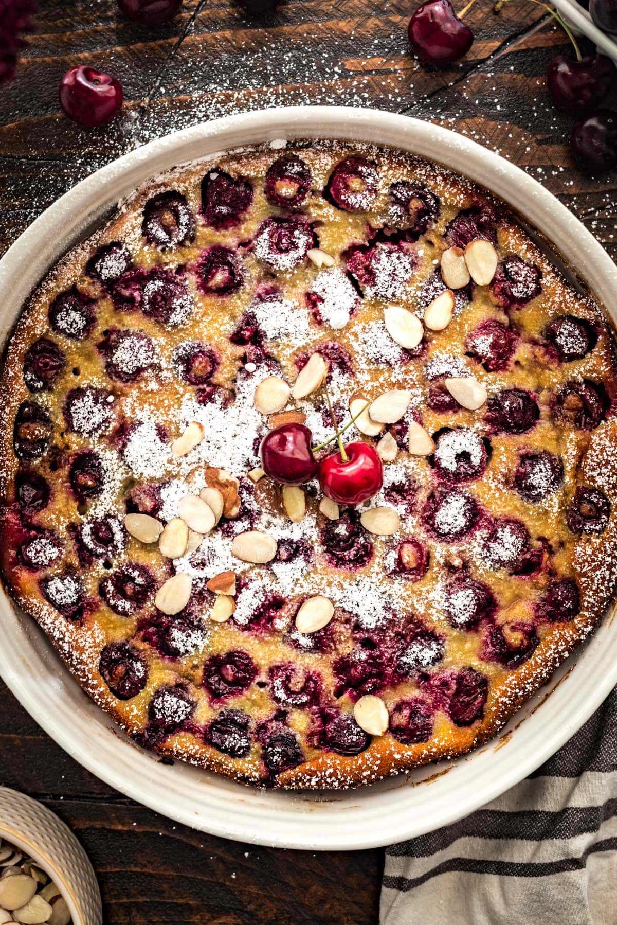 A large cherry clafloutis in a white round pan, and with 2 bright fresh cherries, a dusting of powdered sugar, and toasted sliced almonds on top.