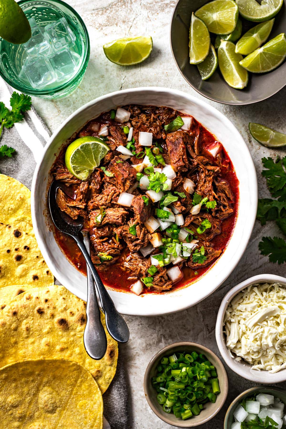 A bowl of birria de res surrounded by oaxaca cheese, limes, and toasted corn tortillas