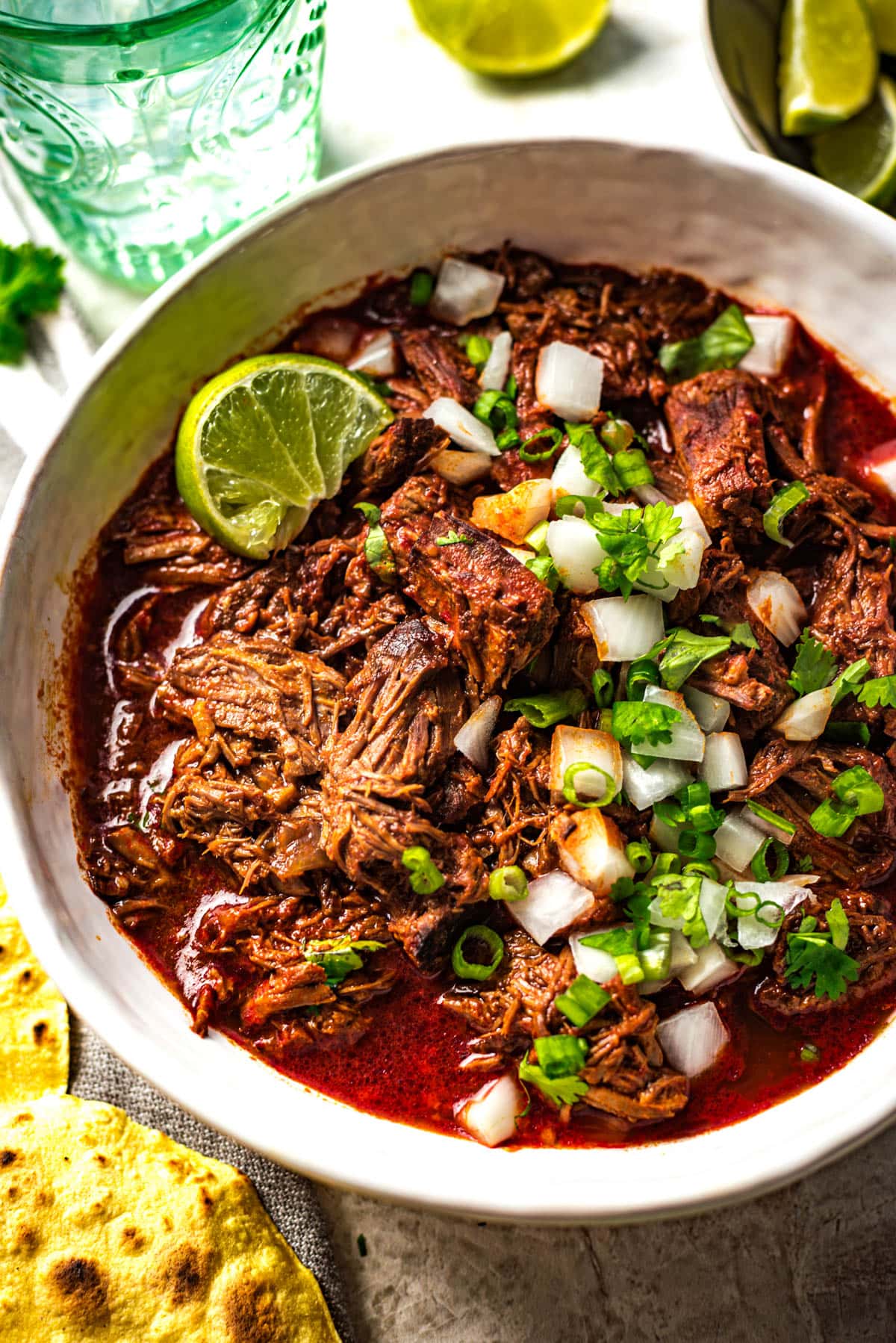 Close up of a bowl filled with beef birria stew, topped with chopped onion, cilantro, green onions, and lime.