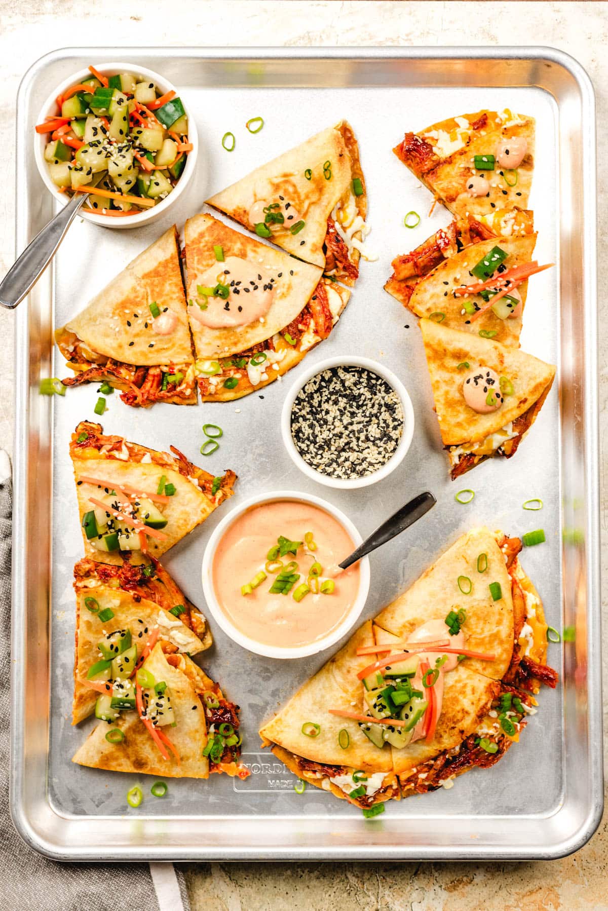 A sheet pan topped with prepared Korean fusion quesadillas, loaded up with gochujang glazed chicken and melted cheese. The quesadillas are sliced and served with gochujang crema, quick pickled vegetables, and sesame seeds.